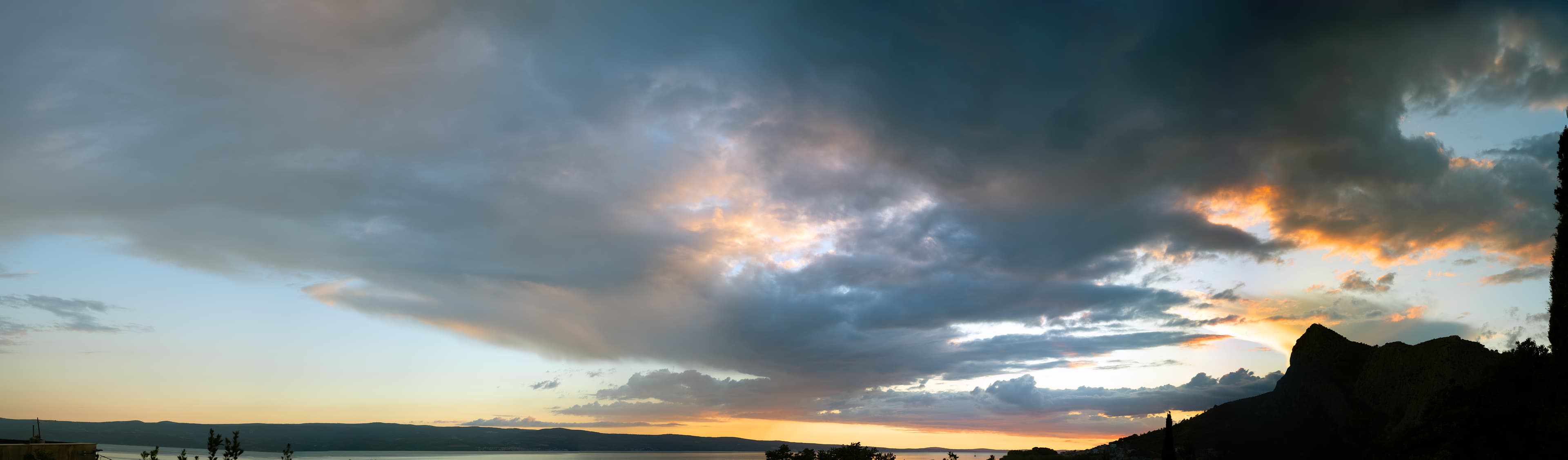 Panoramic sky over the Croatian coast, heavy clouds parting to a glowing golden gap above calm water.