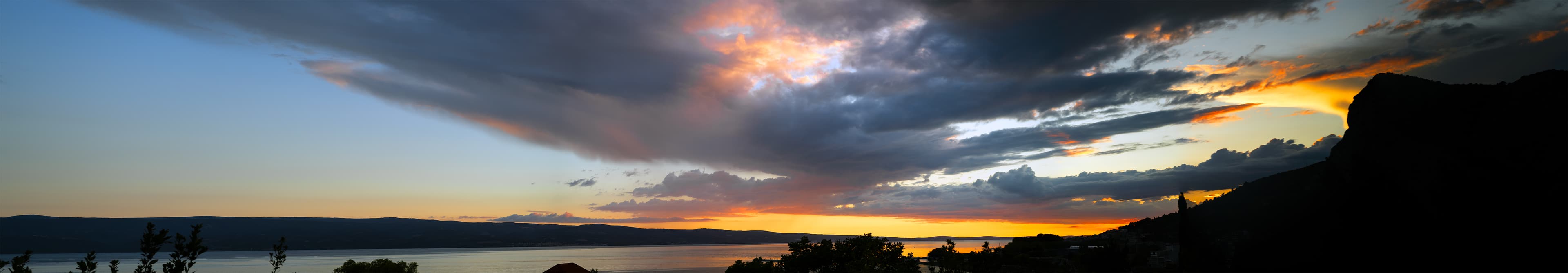Wide panorama of storm clouds breaking into orange and pink light over the Adriatic Sea at sunset.