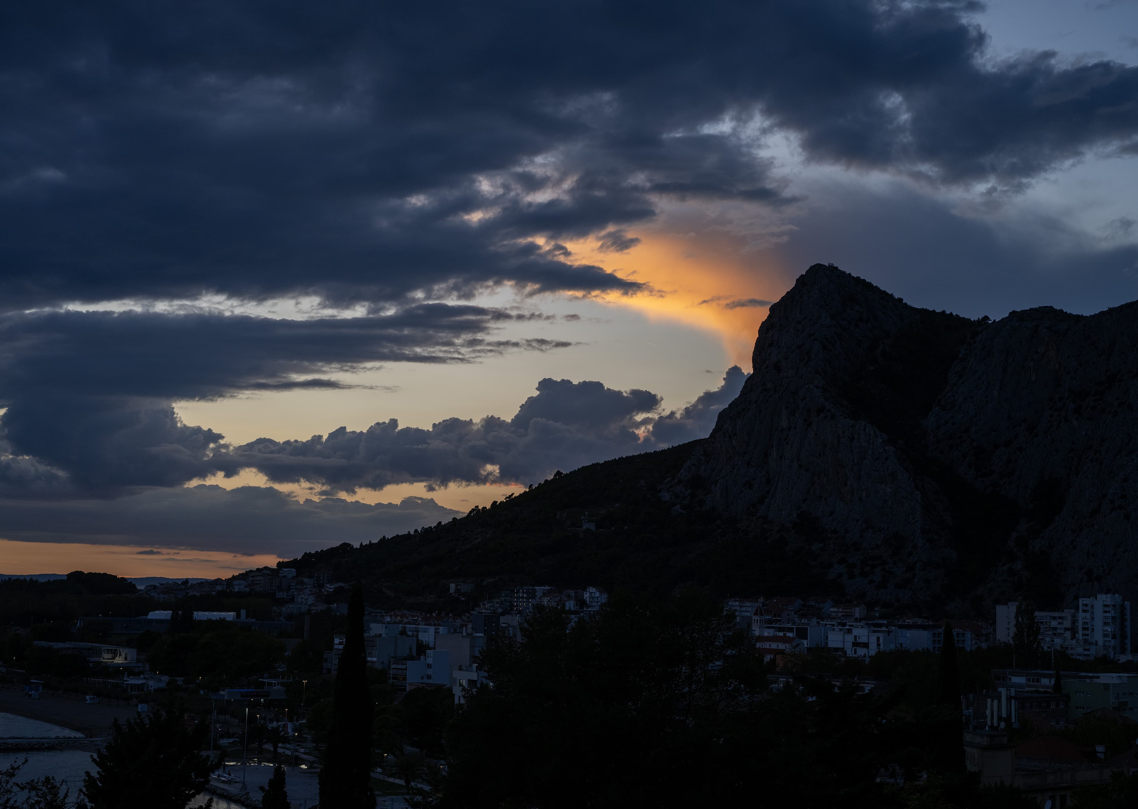 A coastal mountain silhouetted against a stormy sunset sky, with town lights glowing at its base.