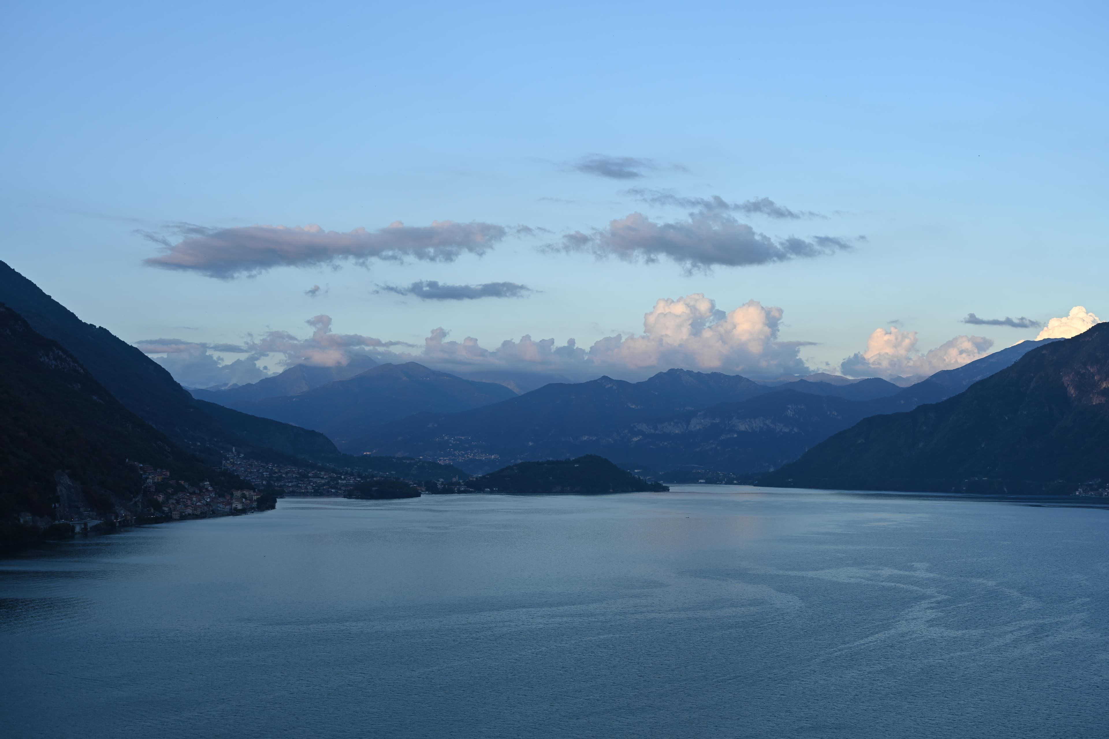 Lake Como at dusk, still blue water framed by layered mountain ridges under a softly clouded sky.