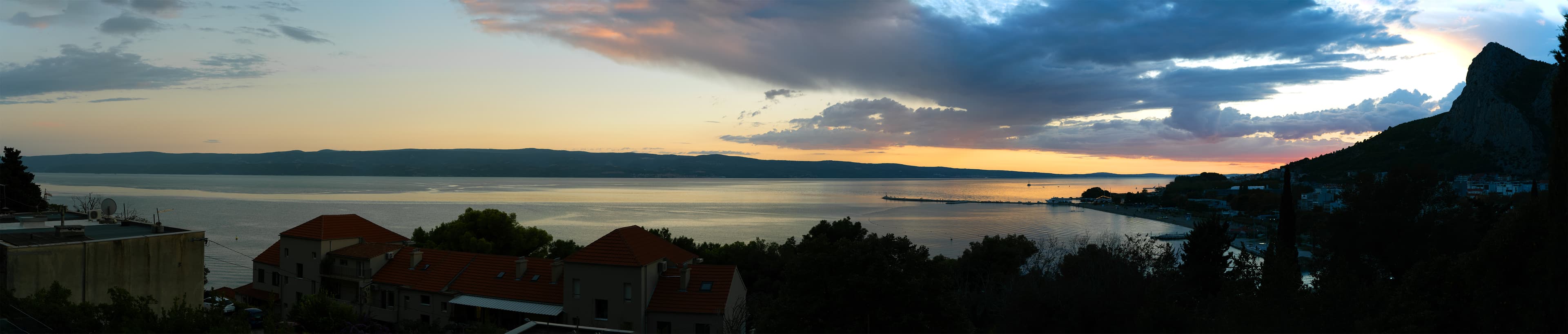 Panoramic view of a red-roofed Croatian coastal village at dusk, with a dramatic mountain silhouette rising over the bay.