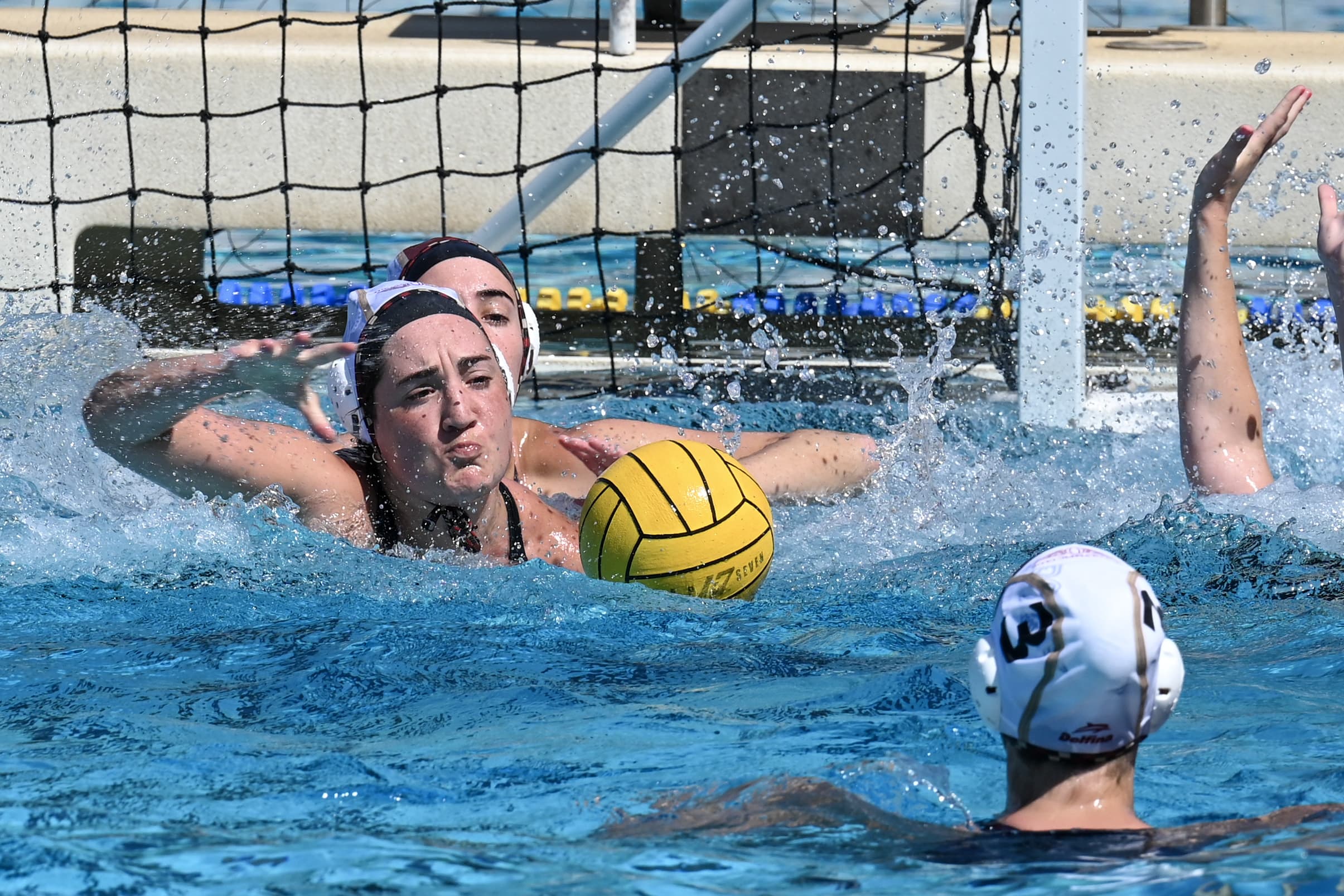 Water polo players contesting a shot at the goal, the ball airborne between them.