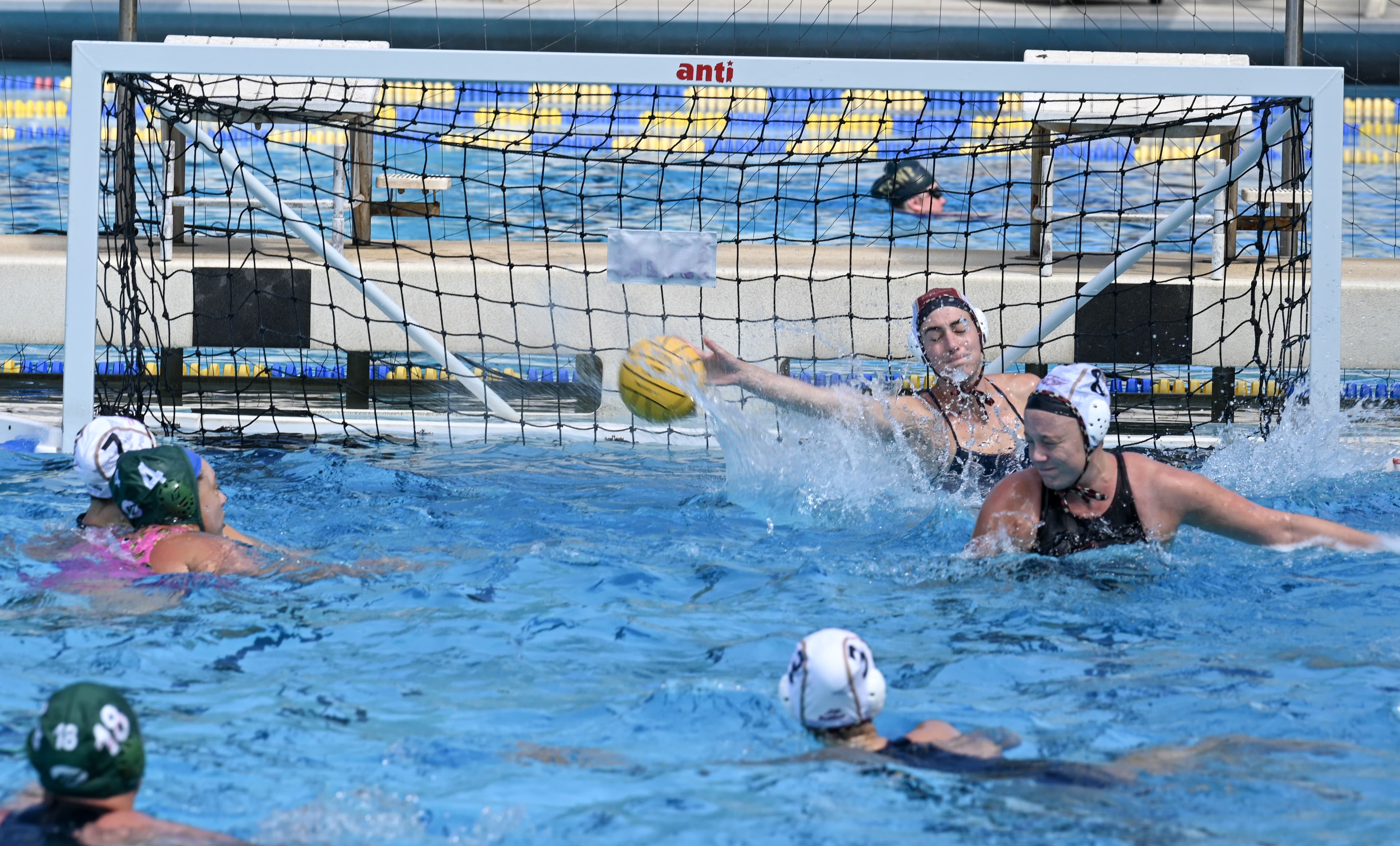 A wide view of a water polo goal scramble, players in different colored caps reaching for the ball near the net.