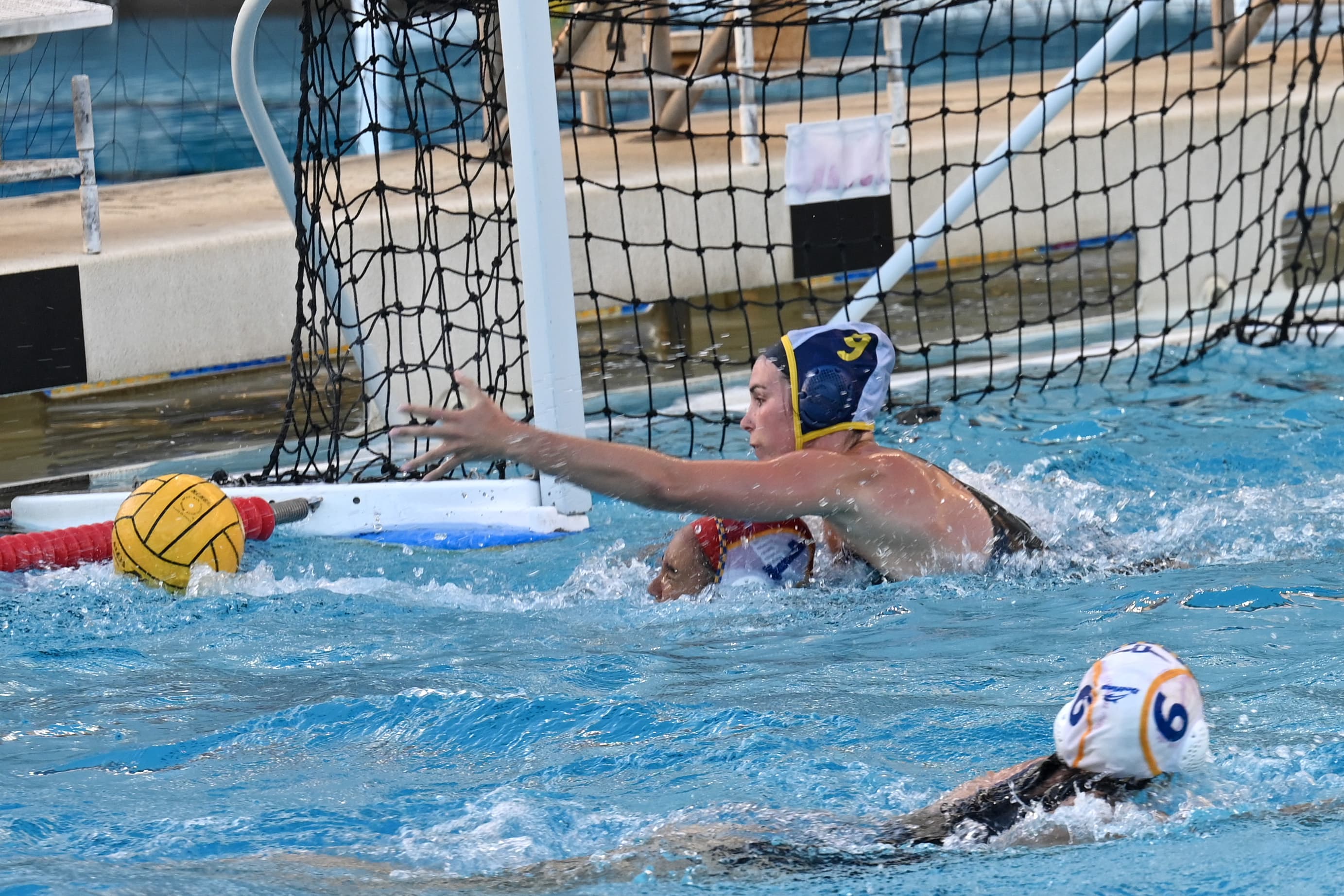 A water polo player in a red cap reaching up for the ball near the goal.